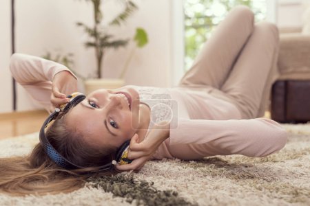 Téléchargez les photos : Belle brune allongée sur le tapis sur le sol de son appartement, écoutant la musique et profitant de son temps libre - en image libre de droit