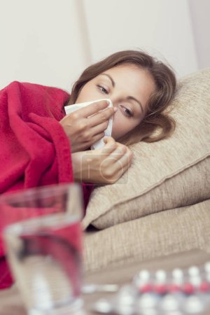 Téléchargez les photos : Femme malade couverte d'une couverture couchée dans son lit avec une forte fièvre et une grippe, se mouchant. Pilules et verre d'eau sur la table - en image libre de droit