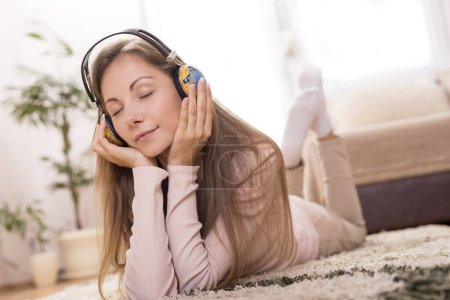 Téléchargez les photos : Belle brune allongée sur le tapis sur le sol de son appartement, écoutant la musique et profitant de son temps libre - en image libre de droit