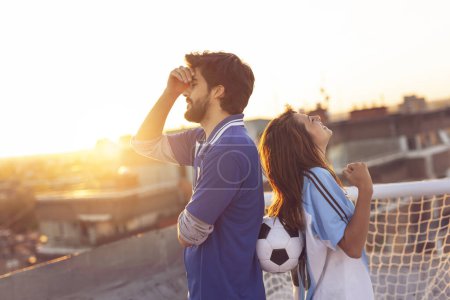 Téléchargez les photos : Couple amoureux portant des maillots de football, debout sur un toit de bâtiment après un match, fille heureuse de célébrer parce que son équipe a gagné, gars triste - en image libre de droit