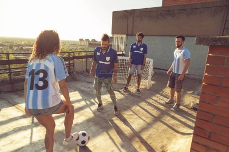 Foto de Grupo de jóvenes amigos con camisetas que se divierten jugando al fútbol en una terraza en la azotea de un edificio en un día soleado de verano - Imagen libre de derechos
