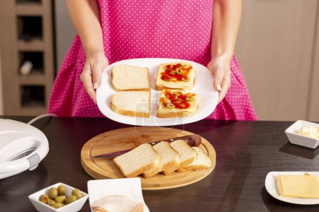 Foto de Mujer sosteniendo un plato con sándwiches listos para calentar en una sandwichera; mujer haciendo sándwiches calientes para el desayuno - Imagen libre de derechos