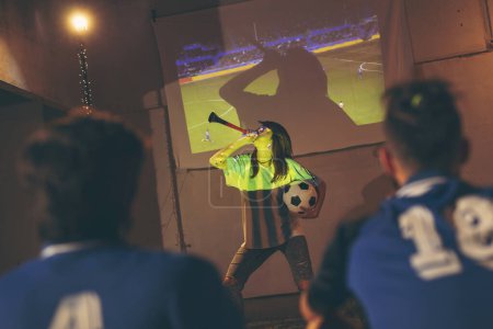 Foto de Grupo de jóvenes amigos viendo un partido de fútbol en la azotea de un edificio; chica sosteniendo un balón de fútbol y tocando la trompeta después de que su equipo anotara un gol - Imagen libre de derechos