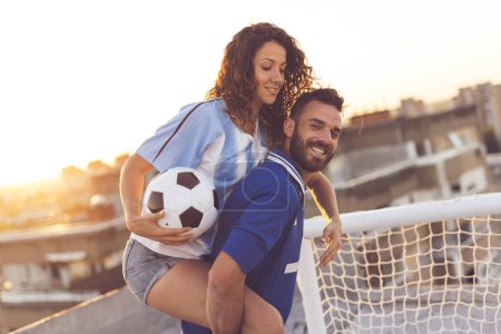 Téléchargez les photos : Jeune couple amoureux s'amuser sur un toit de bâtiment après avoir joué au football. Le gars qui fait un tour en dos de porc à sa copine - en image libre de droit