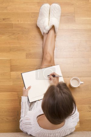 Téléchargez les photos : Vue du dessus d'une jeune femme tenant un cahier, prenant une tasse de café et écrivant dans son journal, appréciant dans une atmosphère de maison confortable - en image libre de droit