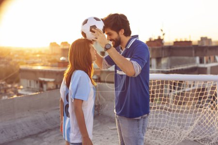 Téléchargez les photos : Jeune couple amoureux s'amuser sur un toit de bâtiment après avoir joué au football, danser et équilibrer avec le ballon entre leurs têtes - en image libre de droit