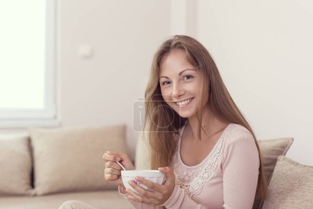 Photo for Young woman sitting on a living room couch, holding a bowl of cereal and having breakfast - Royalty Free Image