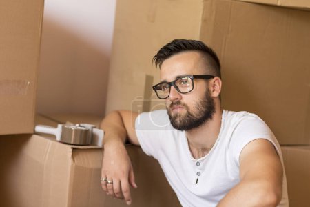 Foto de Joven hombre que se mueve en un nuevo apartamento, sentado en el suelo, rodeado de cajas de cartón con máquina de embalaje colocado en una de las cajas en el fondo - Imagen libre de derechos