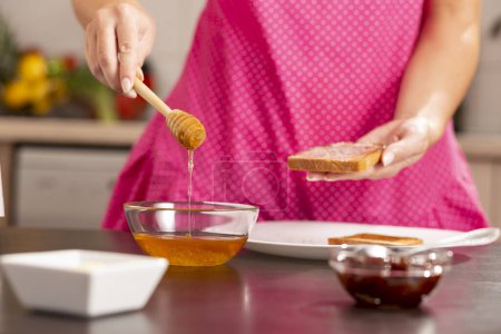 Téléchargez les photos : Détail des mains féminines tenant une tranche de pain grillé et une cuillère en bois, étalant le miel sur le pain ; Concentration sélective sur la cuillère et le miel qui fuit - en image libre de droit
