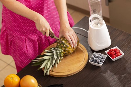 Téléchargez les photos : Vue en angle élevé d'une femme coupant un dessus d'ananas avec un couteau de cuisine sur une planche à découper afin de le peler avec un coupe-ananas. Focus sur le couteau - en image libre de droit