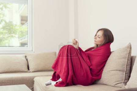 Photo for Young woman sitting on a couch covered with blanket, having a fever, holding a thermometer after measuring temperature. - Royalty Free Image