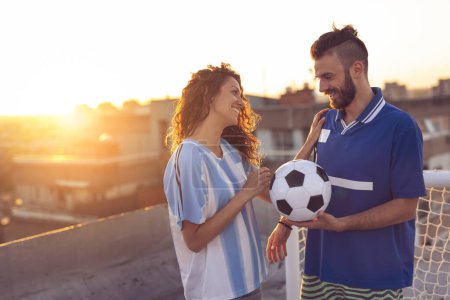 Foto de Pareja enamorada vistiendo camisetas de fútbol, de pie en una azotea del edificio después de un partido y disfrutando de una hermosa puesta de sol sobre la ciudad. Concéntrate en la chica - Imagen libre de derechos