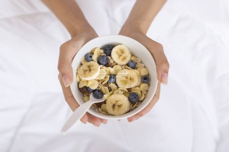 Photo for Close up of a young woman's hands holding a bowl of breakfast cereal mix with blueberries and bananas. Selective focus - Royalty Free Image