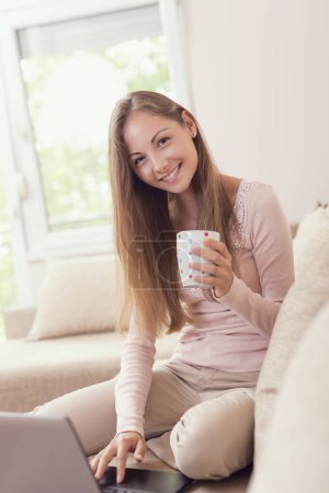 Téléchargez les photos : Belle jeune brune assise sur un canapé salon, tenant une tasse de café et utilisant un ordinateur portable - en image libre de droit