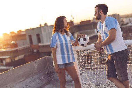 Foto de Pareja enamorada vistiendo camisetas de fútbol, de pie en la azotea de un edificio después de un partido y disfrutando de una hermosa puesta de sol sobre la ciudad - Imagen libre de derechos