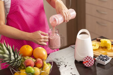 Photo for Detail of female hands pouring a freshly made raspberry smoothie in a jar; woman wearing apron standing in the kitchen, making smoothie for breakfast - Royalty Free Image