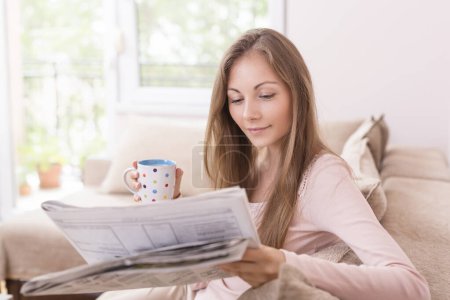 Photo for Young beautiful woman sitting on the couch in the living room, reading the newspapers and drinking coffee - Royalty Free Image