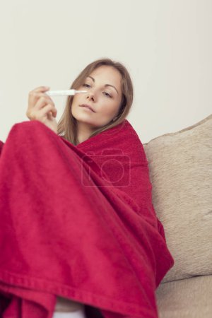 Foto de Mujer joven sentada en un sofá cubierto de manta, con fiebre, sosteniendo un termómetro después de medir la temperatura. - Imagen libre de derechos