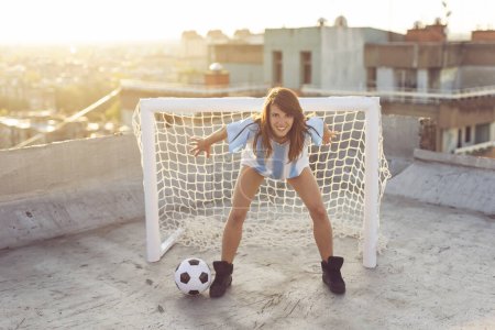 Photo for Beautiful young female football fan standing in front of a goal as a goalkeeper on a building rooftop terrace, having fun while waiting for a match to start - Royalty Free Image
