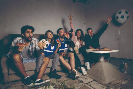 Foto de Grupo de jóvenes amigos viendo un partido de fútbol en la azotea de un edificio, animando a comer pizza y beber cerveza, alegres después de anotar un gol - Imagen libre de derechos