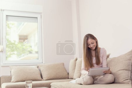 Foto de Hermosa joven sentada en un sofá de la sala de estar, sosteniendo una tableta y navegar por la red - Imagen libre de derechos