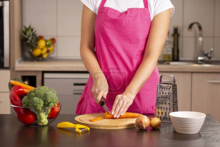Téléchargez les photos : Détail des mains féminines coupant des carottes fraîches avec un couteau de cuisine sur une planche à découper pour faire de la salade de légumes frais - en image libre de droit