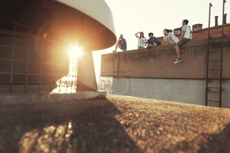 Foto de Grupo de jóvenes amigos que se divierten jugando al fútbol en la azotea de un edificio, tomando un descanso y disfrutando de la puesta de sol sobre la ciudad. Centrarse en la gente en el medio - Imagen libre de derechos