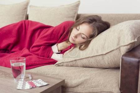Téléchargez les photos : Femme malade couverte d'une couverture couchée dans son lit avec une forte fièvre et une grippe. Pilules et verre d'eau sur la table - en image libre de droit
