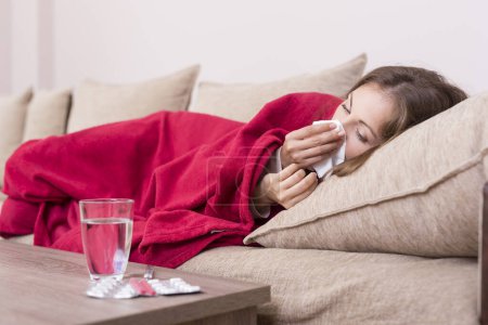 Foto de Mujer enferma cubierta con una manta acostada en la cama con fiebre alta y gripe, sonándose la nariz. Pastillas y vaso de agua sobre la mesa - Imagen libre de derechos