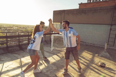 Foto de Grupo de jóvenes amigos divirtiéndose jugando al fútbol en una terraza de la azotea del edificio, equipo ganador celebrando anotar un gol, haciendo cinco - Imagen libre de derechos