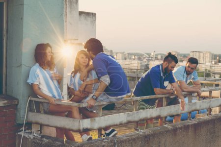 Groupe de fans de football debout sur un toit-terrasse du bâtiment, traînant en attendant le début du match ; amis s'amusant à regarder le football se détendre pendant une mi-temps