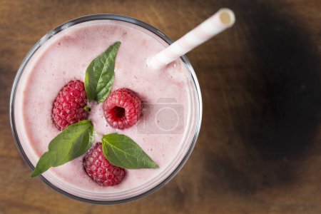 Photo for Top view of a raspberry smoothie glass, decorated with drinking straw, three raspberries and mint leaves, placed on a rustic wooden cutting board - Royalty Free Image