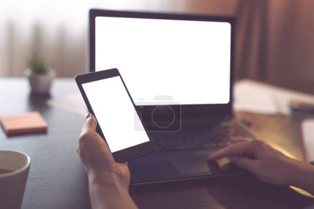 Photo for Mockup image of female hands using a laptop computer and holing a smart phone both with blank white screens. Focus on the left hand's thumb - Royalty Free Image