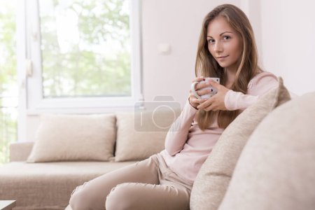 Téléchargez les photos : Belle jeune femme assise sur le canapé du salon, tenant une tasse de café - en image libre de droit