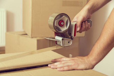 Photo for Close up of a guy's hands holding packing machine and sealing cardboard boxes with duct tape - Royalty Free Image