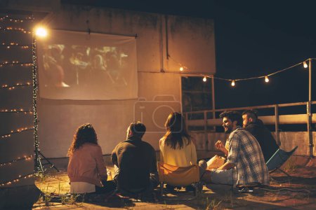 Téléchargez les photos : Groupe de jeunes amis regardant un film sur un toit-terrasse, mangeant du pop-corn, buvant de la bière et s'amusant - en image libre de droit