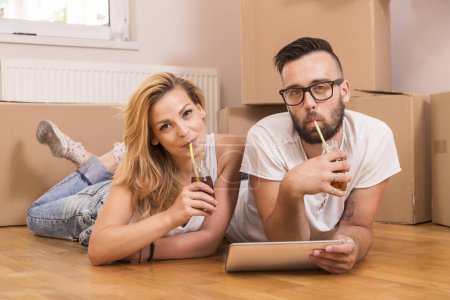 Téléchargez les photos : Jeune couple amoureux se déplaçant dans un nouvel appartement, couché sur le sol et surfant sur le web sur une tablette informatique à la recherche de nouvelles idées de redécoration - en image libre de droit