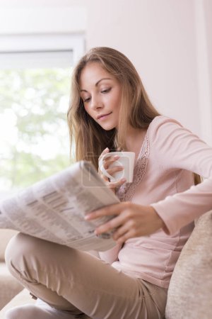Téléchargez les photos : Jeune belle femme assise sur le canapé dans le salon, lisant les journaux et buvant du café - en image libre de droit