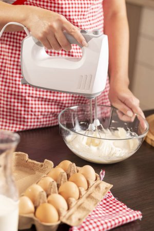 Téléchargez les photos : Détail des mains féminines tenant le mélangeur et mélangeant la pâte à crêpes sur le comptoir de la cuisine. Concentrez-vous sur le mélangeur et la main qui le tient - en image libre de droit