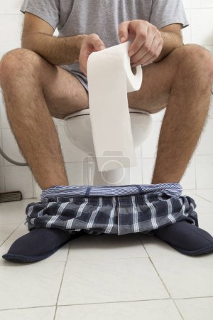 Téléchargez les photos : Détail d'un homme assis sur les toilettes avec son pyjama et ses sous-vêtements, tenant un rouleau de papier toilette - en image libre de droit