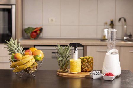 Photo for Fresh fruits in a bowl and a pineapple peeled with pineapple cutter on a kitchen counter next to a smoothie maker - Royalty Free Image