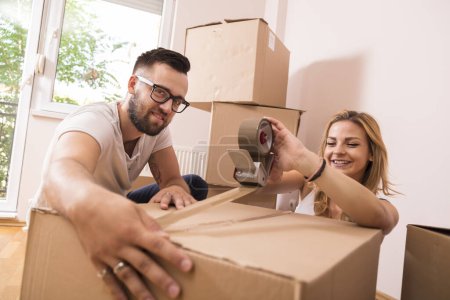 Photo for Young girl moving in a new apartment with her boyfriend,standing surrounded with cardboard boxes, packing and taping boxes, while the boyfriend carries boxes away - Royalty Free Image