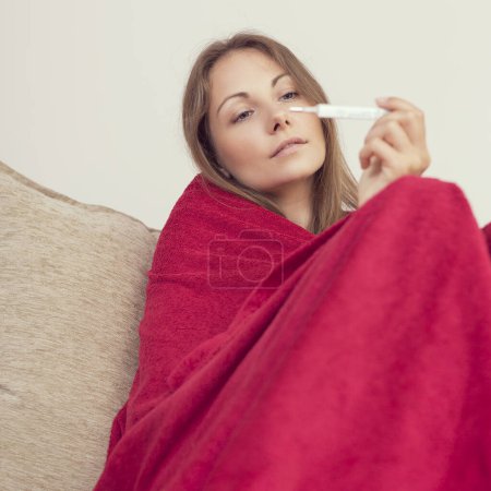 Foto de Mujer joven sentada en un sofá cubierto de manta, con fiebre, sosteniendo un termómetro después de medir la temperatura. - Imagen libre de derechos
