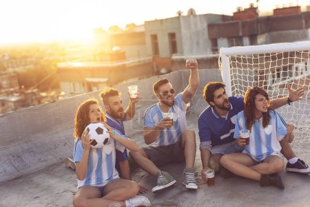 Foto de Jóvenes amigos viendo un partido de fútbol, animando a su equipo y bebiendo cerveza en la azotea de un edificio, con paisaje urbano en el fondo - Imagen libre de derechos