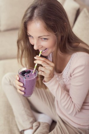 Photo for Beautiful girl sitting on a living room couch, smiling and holding a glass of raspberry and blueberry mix smoothie. - Royalty Free Image