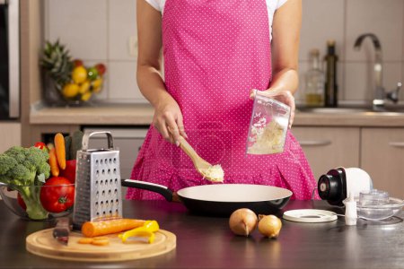 Téléchargez les photos : Détail des mains féminines mettant l'oignon haché dans une poêle avec une cuillère de cuisine à partir d'un bol à oignon sur le comptoir de la cuisine - en image libre de droit