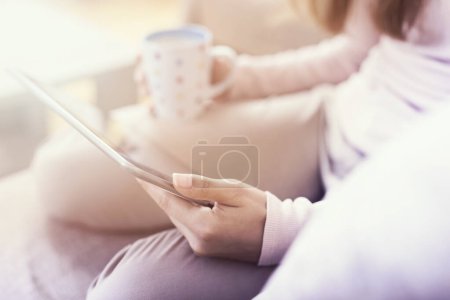 Téléchargez les photos : Détail d'une belle jeune fille assise sur un canapé du salon, tenant une tablette, une tasse de café et surfant sur le net - en image libre de droit