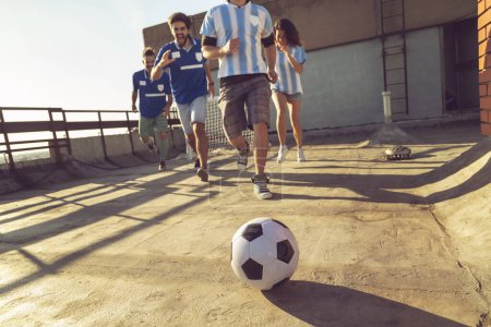 Foto de Grupo de jóvenes amigos vistiendo camisetas divirtiéndose jugando al fútbol en una terraza en la azotea de un edificio en un día soleado de verano, todos corriendo hacia la pelota - Imagen libre de derechos