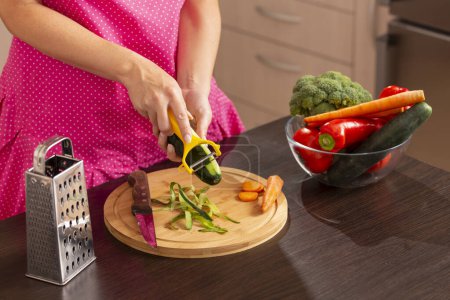 Foto de Detalle de las manos femeninas utilizando una rebanadora de pepino para pelar un pepino para hacer una ensalada de verduras frescas - Imagen libre de derechos