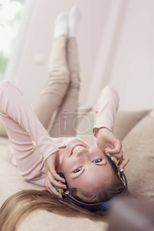 Téléchargez les photos : Jeune femme séduisante allongée sur un canapé dans le salon, portant un casque, écoutant la musique et profitant de sa liberté et de ses loisirs - en image libre de droit